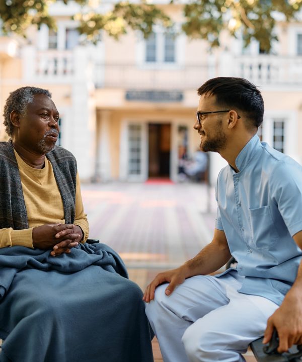 Black senior in wheelchair talking to male nurse while relaxing in park of residential care home.